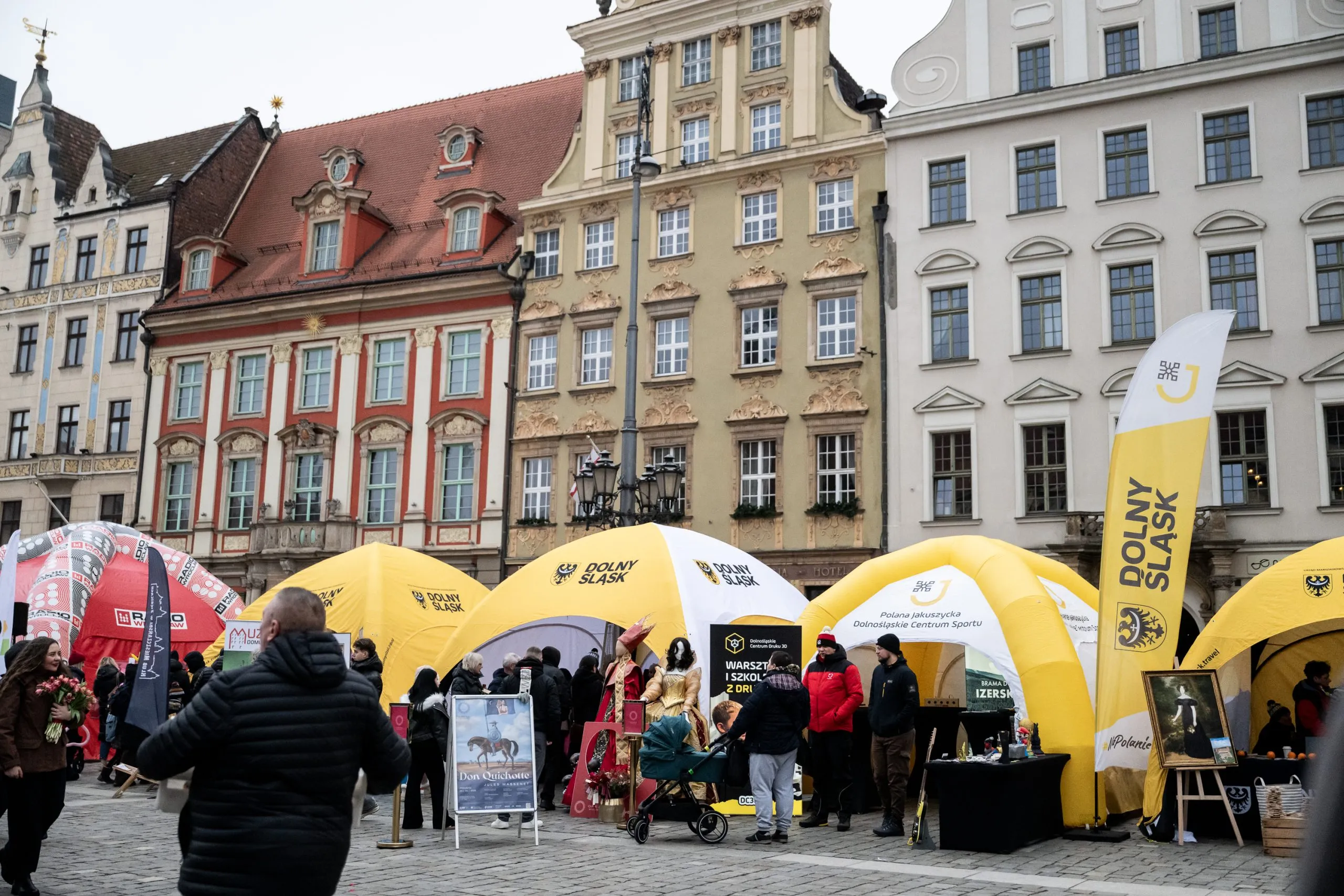 Dolnośląskie stoisko promocyjne z żółtymi namiotami i flagami przyciąga odwiedzających na zabytkowym rynku otoczonym kolorowymi kamienicami.