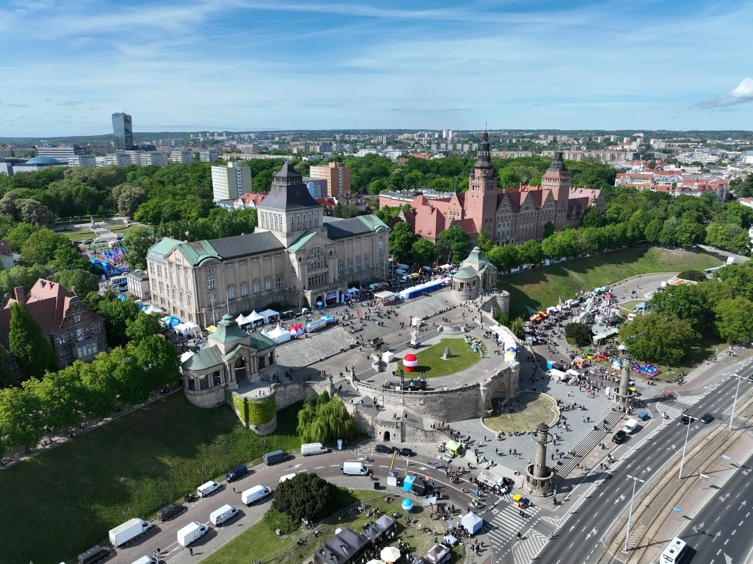 Widok z lotu ptaka na rozległy plac miejski przygotowany do plenerowego wydarzenia, z budynkami, zielonym parkiem, stoiskami i grupami odwiedzających.