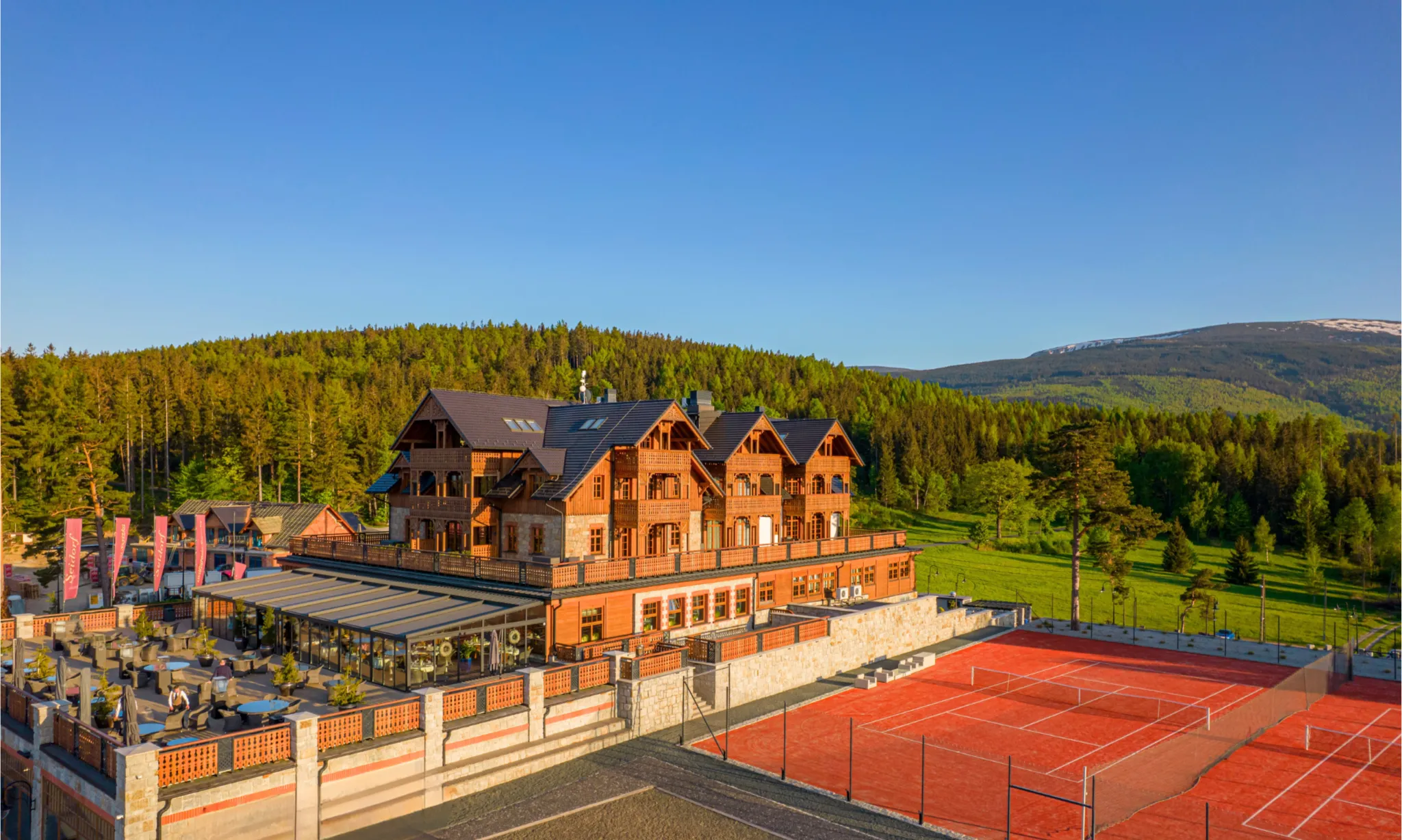 Elegancki hotel w stylu górskiego kurortu, położony wśród zielonych lasów u podnóża gór. Obok budynku znajduje się kort tenisowy oraz przestronny taras, a całość otacza malowniczy krajobraz skąpany w ciepłym świetle.