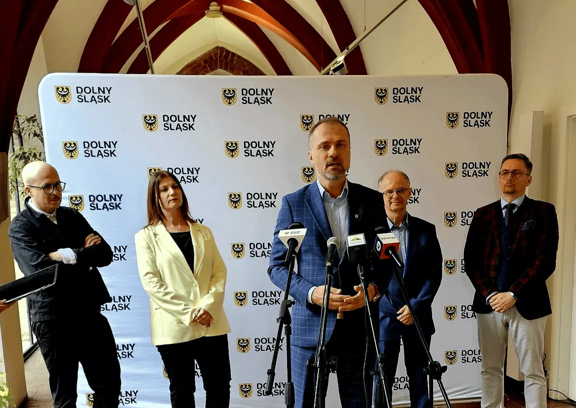 Pięć osób siedzi za stołem podczas konferencji prasowej, przed mikrofonami i ścianką z logotypami instytucji kultury oraz konkursu muzealnego.