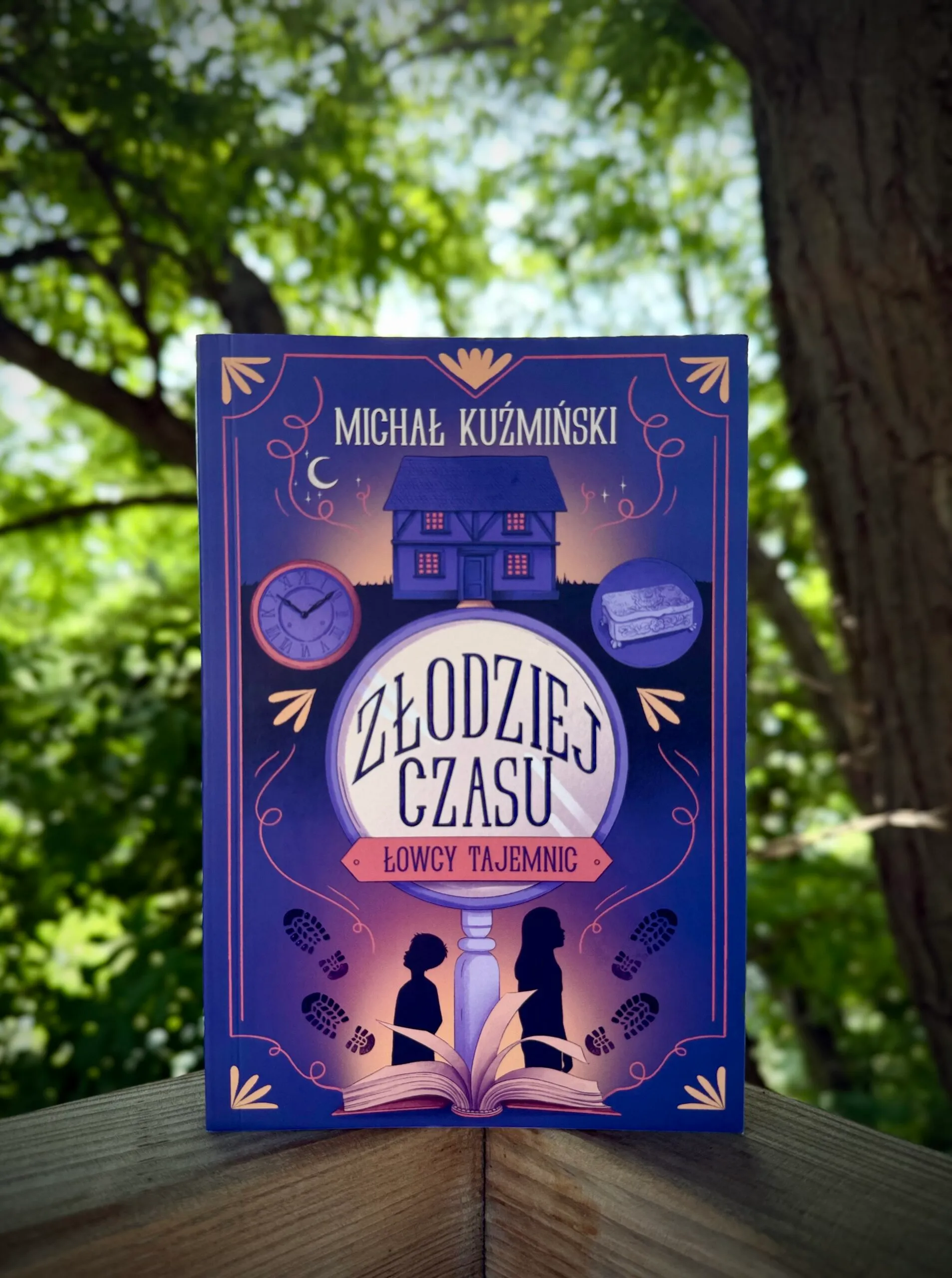 Książka „Złodziej czasu” Michaiła Kostrowickiego stoi na drewnianym stole wśród zieleni, eksponując fioletową okładkę z magicznym zegarem i otwartą księgą.
